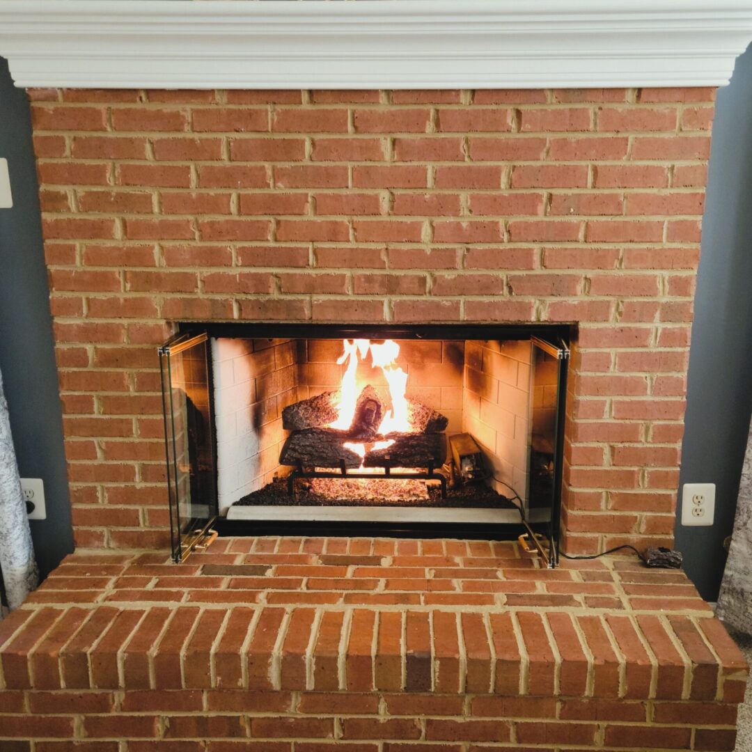 brick fireplace with the doors open that show a burning gas log set. There is a white mantle above the fireplace opening.